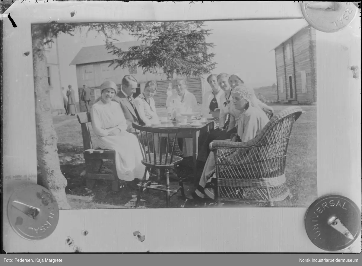 Reproduksjonsfotografi av eldre positiv.Gruppe mennesker sittende rundt born på gårdsplass. To laftede bygg i bakgrunnen.