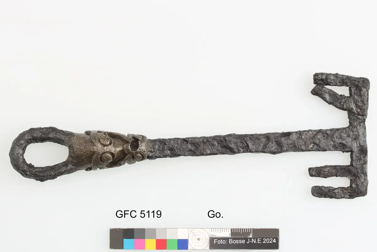 Huvudet består av en ögla nedanför denna pålagd bronsornamentik i form av växtornamentik stammen massiv med rektangulärt tvärsnitt dubbelsidigt ax enligt orginalkatalogen funnen under plöjning på ca 25 cm djup omkring 300 m från boningshuset
av järn, med bronsbeslag. Längd 167,5 mm. Från boticke i anga. Funnen under plöjning på 2,5 dm. Djup, omkring 300 meter från boningshuset. Enligt uppgift av a. Jansson.