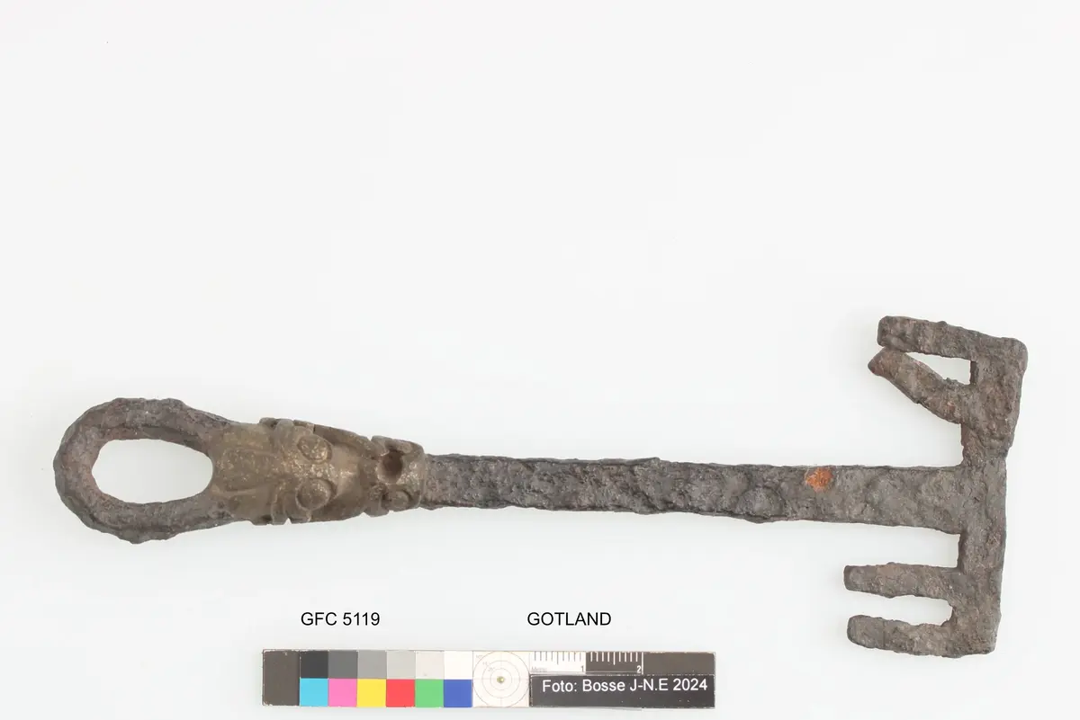 Huvudet består av en ögla nedanför denna pålagd bronsornamentik i form av växtornamentik stammen massiv med rektangulärt tvärsnitt dubbelsidigt ax enligt orginalkatalogen funnen under plöjning på ca 25 cm djup omkring 300 m från boningshuset
av järn, med bronsbeslag. Längd 167,5 mm. Från boticke i anga. Funnen under plöjning på 2,5 dm. Djup, omkring 300 meter från boningshuset. Enligt uppgift av a. Jansson.