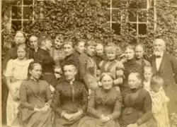 Stor samling av folk foran en eføybekledd vegg. Før 1897.  K