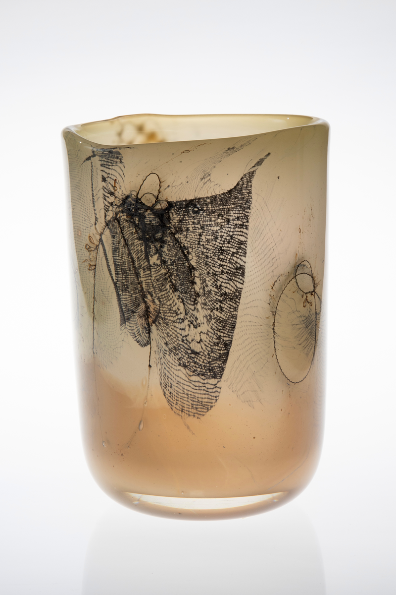 Sylinderformet vase i halvgjennomskinnelig glass. Ujevn munning. Grågrønt underfang som er dekorert med innlagte metalltråder samt partier med finmasket blålig netting. Tidvis brune spetter samt luftbobler i glassmassen. Puntemerke under vasens bunn.