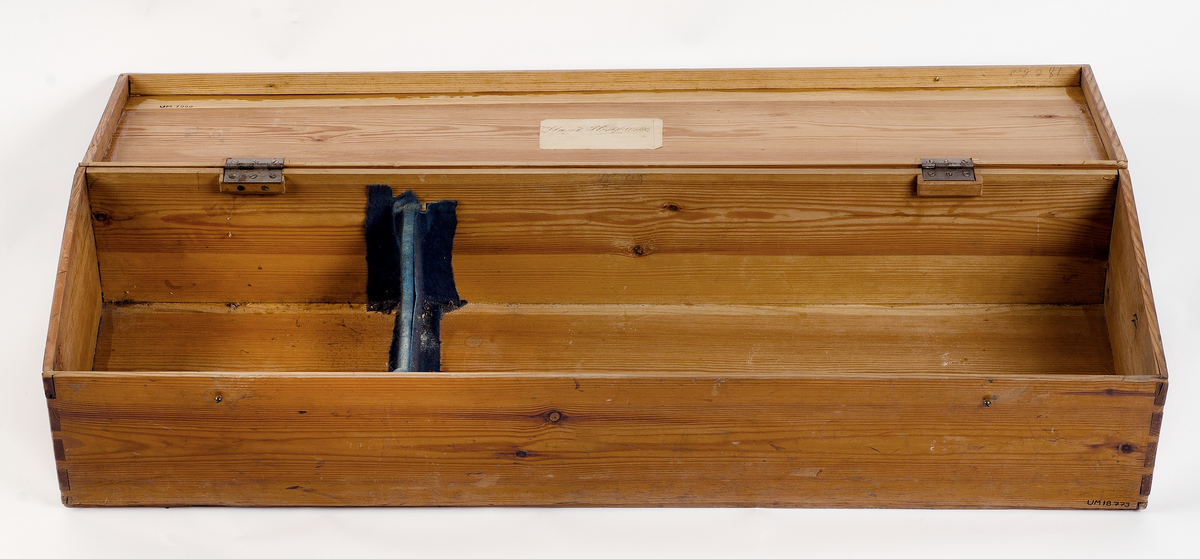 Förvaringslåda för nyckelharpa. Tillverkad av furu med zinkade hörn. Bärhandtag av gulmetall och låskläppar av gulmetall. Inne i lådan ett stöd för harpan klätt med blå filt. Påklistrad etikett med ägarens namn: Knut Håkansson.
