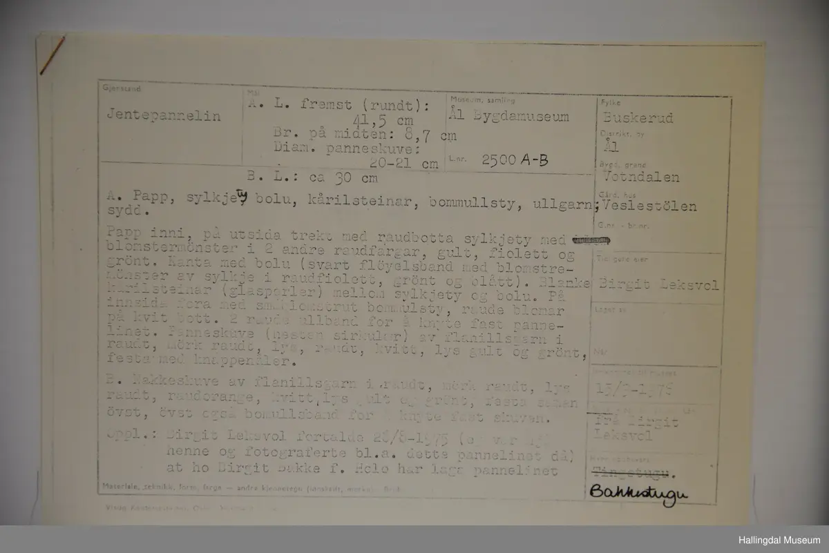 a) Jentepannelin av papp, sylkjety, bolu, kårilsteinar, bomullsty, ullgarn, sydd.
b) Nakkeskuve av flanillsgarn i raudt, mørk raudt, lys raudt, kvitt, lys gult, og grønt.