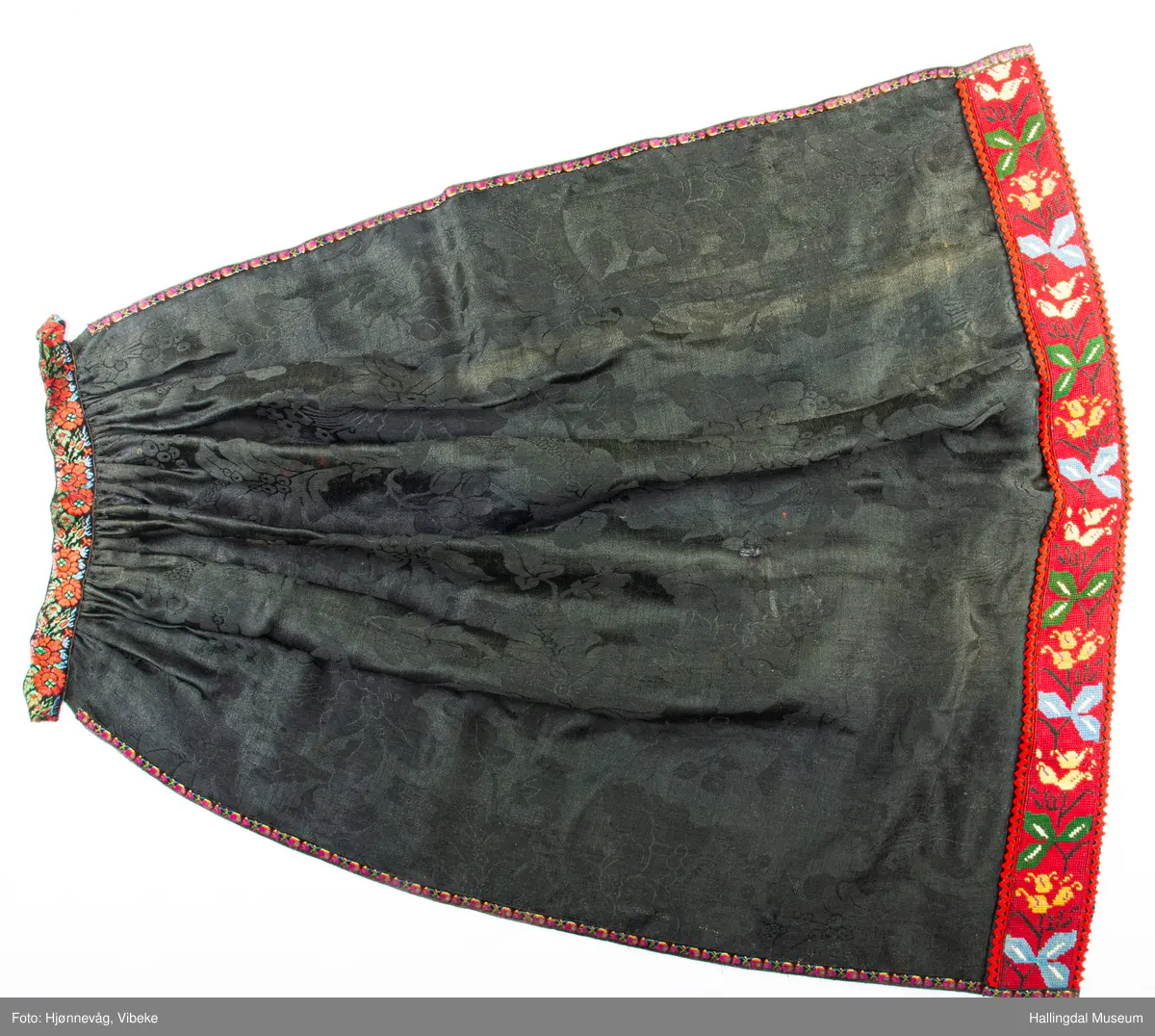 Damask med band sauma på stramei. Sydd og sauma. Rektangulært tøystykke rynka øvst. Ei brei bole øverst, smale boler på sidene. Lengde: 110 cm, bredde: 94 cm, 