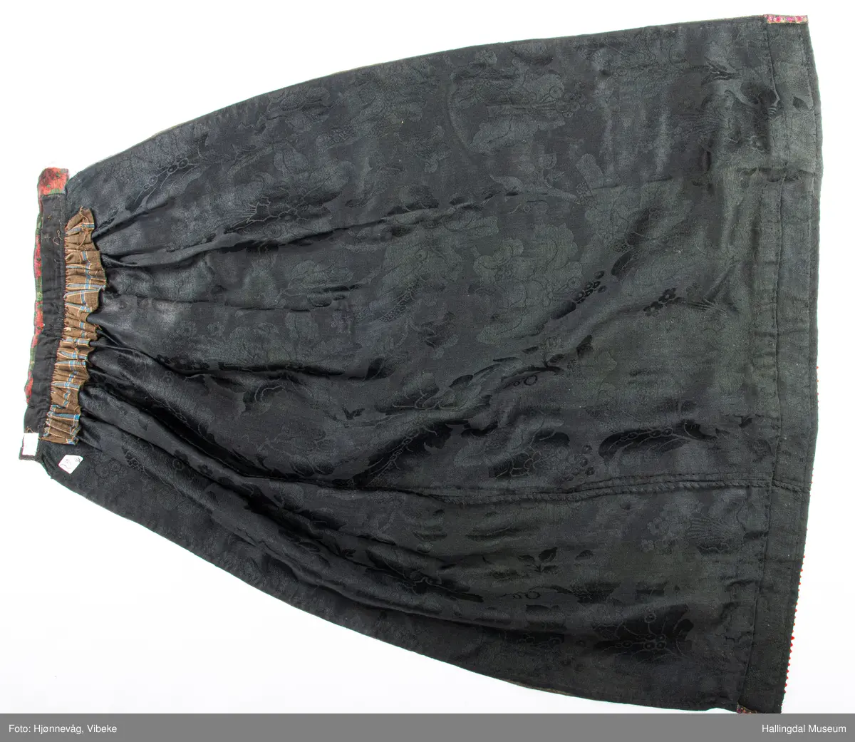 Damask med band sauma på stramei. Sydd og sauma. Rektangulært tøystykke rynka øvst. Ei brei bole øverst, smale boler på sidene. Lengde: 110 cm, bredde: 94 cm, 
