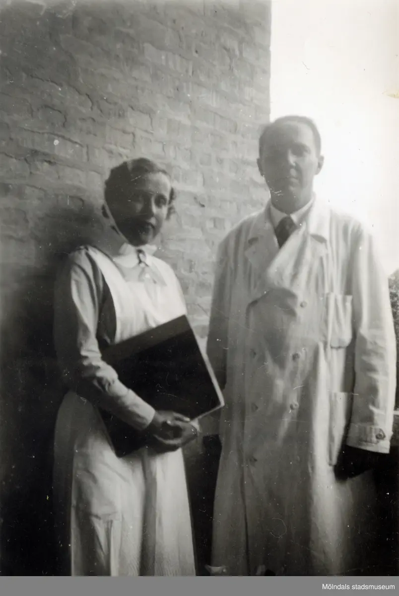 En sjuksköterska och en läkare står vid en tegelvägg utanför Mölndals sjukhus 1939 - 1941. Bilder från fotoalbum, tagna från södra sidan av Länsmansgatan vid Mölndals lasarett/sjukhus 1939 - 1941 på bland annat givarens mor Ester (född Johansson) tillsammans med kollegan/väninnan Aina Englund. De arbetade på den manliga kirurgavdelningen som vårdbiträden/undersköterskor.
Relaterade motiv: 2024_1542 - 1617.