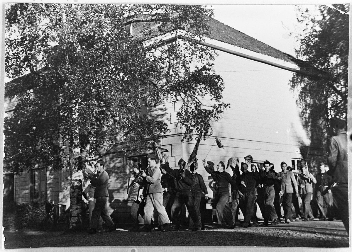 Fra AT-leiren på Kjølseth gård på Skreia 17.mai 1943. AT-mannskapene har laget demonstrsjonstog i anledning nasjonaldagen, med musikk og ei fille på en stav som erstatning for flagg. Dette var selvsagt forbudt, men ettersom de var så mange ble det ikke så mange represalier. Håkon Andersen mener å huske at det ble en del permisjonsnekt.