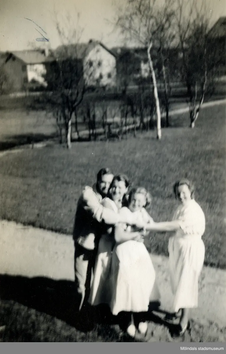 Tre sjukvårdsbiträden och en manlig patient står på en gräsmatta och håller i varandra, Mölndals sjukhus 1939 - 1941. Från vänster ses Ester Johansson (1914 - 2008, gift Falkdahl) och Aina Englund (1914 - 2012). I bakgrunden ses några låga bostadshus. Bilder från fotoalbum vid Mölndals lasarett/sjukhus 1939 - 1941 på bland annat givarens mor Ester (född Johansson) tillsammans med kollegan/väninnan Aina Englund. De arbetade på den manliga kirurgavdelningen som vårdbiträden/undersköterskor.
Relaterade motiv: 2024_1542 - 1617.