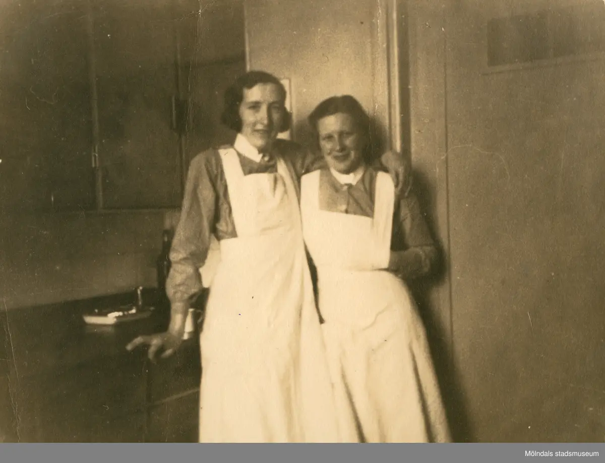 Två kvinnliga sjukvårdsbiträden står framför ett bord och håller om varandra, Mölndals sjukhus 1939 - 1941. Bilder från fotoalbum, tagna från södra sidan av Länsmansgatan vid Mölndals lasarett/sjukhus 1939 - 1941 på bland annat givarens mor Ester (född Johansson) tillsammans med kollegan/väninnan Aina Englund. De arbetade på den manliga kirurgavdelningen som vårdbiträden/undersköterskor.
Relaterade motiv: 2024_1542 - 1617.