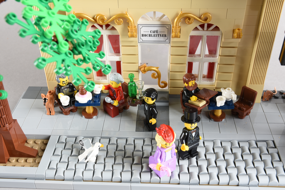 Tablået er bygget av Lego i diverse farger. Scene med syv mennsker stående, gående og sittende utenfor en cafè og bokhandel. Wien 1870, Cafe Hochleitner.