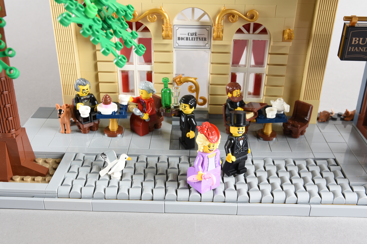 Tablået er bygget av Lego i diverse farger. Scene med syv mennsker stående, gående og sittende utenfor en cafè og bokhandel. Wien 1870, Cafe Hochleitner.