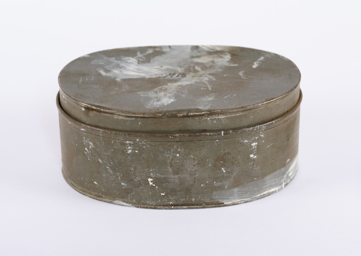 Oval matboks i blikk. Består av to deler, den ene delen går over i den andre. Ring på siden til å bære i. Denne ligger flatt inntil siden.