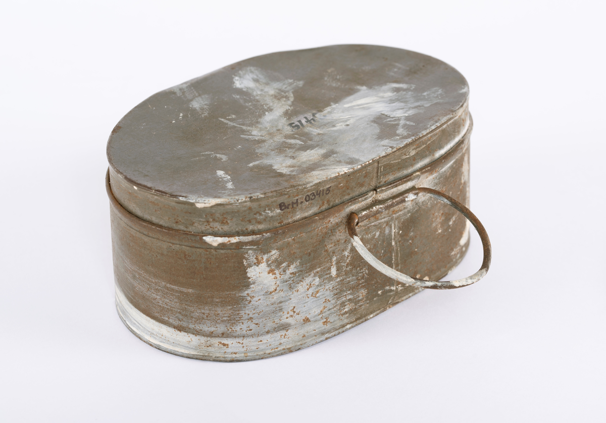 Oval matboks i blikk. Består av to deler, den ene delen går over i den andre. Ring på siden til å bære i. Denne ligger flatt inntil siden.