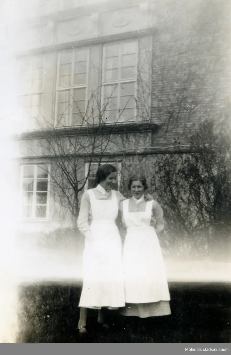 Två sjukvårdsbiträden står på en gräsmatta och håller om varandra, Mölndals sjukhus 1939 - 1941. Ljusskadat foto. Bilder från fotoalbum vid Mölndals lasarett/sjukhus 1939 - 1941 på bland annat givarens mor Ester (född Johansson) tillsammans med kollegan/väninnan Aina Englund. De arbetade på den manliga kirurgavdelningen som vårdbiträden/undersköterskor.
Relaterade motiv: 2024_1542 - 1617.