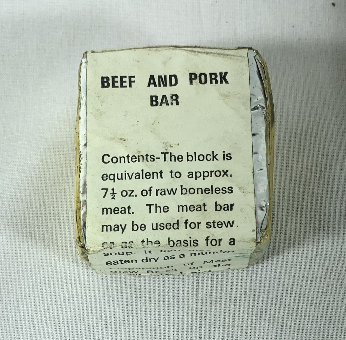 Firkantet blokk merket "Beef and Pork Bar" pakket inn i folie med hvit papiretikett. Foran står det at innholdet tilsvarer omtrent 7 ½ oz. rå benfritt kjøtt, og at kjøttbaren kan brukes til gryteretter eller suppe. På baksiden oppgis ingrediensene som frysetørket, malt okse- og svinekjøtt, spiselig fett, salt og mononatriumglutamat. Instruksjonene nevner å koke kjøttet i vann i fem minutter, med mulighet for å tilsette mer vann under kokingen.














