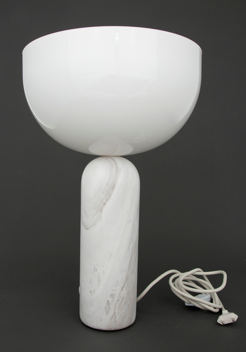 Lampe i dreiet marmor og injeksjonsstøpt akryl. Sylinderformet sokkel i marmor med mykt avrundet øvre del, som møter en halvkuleformet skjerm i akryl.