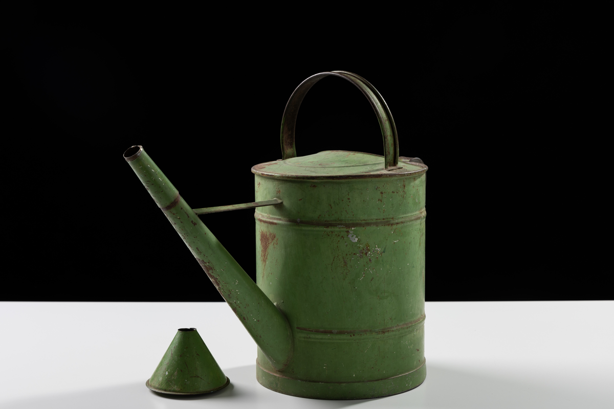 Vattenkanna av metallplåt, grönmålad. Cylinderformad med bågformat handtag upptill och på baksidan. Skvalpskydd och lång pip med lös striltratt.