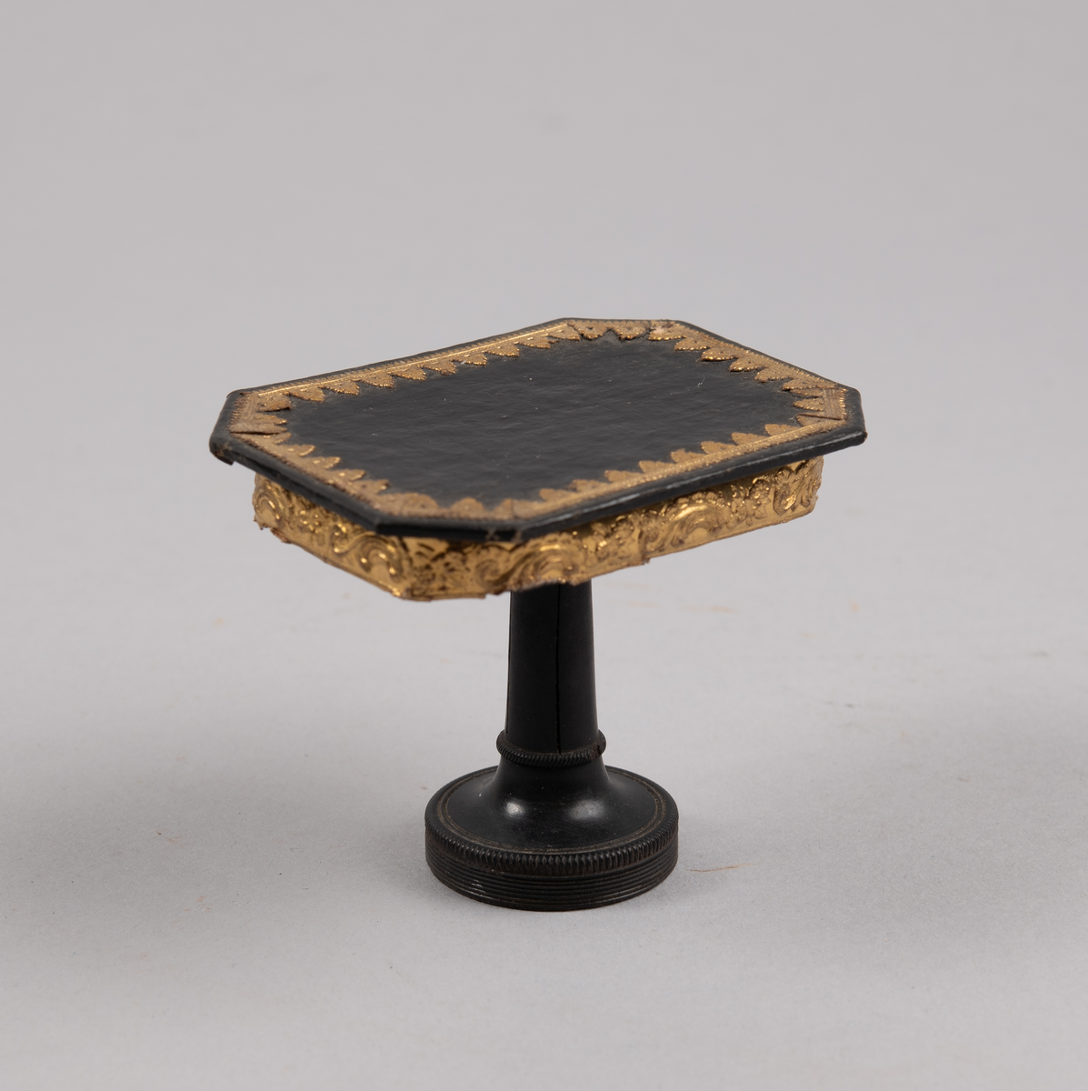 Bord til dukkehus. 8-kantet bordplate med gulldekor langs kanten. En dreid søylefot.