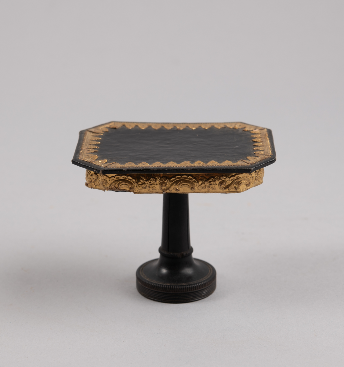 Bord til dukkehus. 8-kantet bordplate med gulldekor langs kanten. En dreid søylefot.