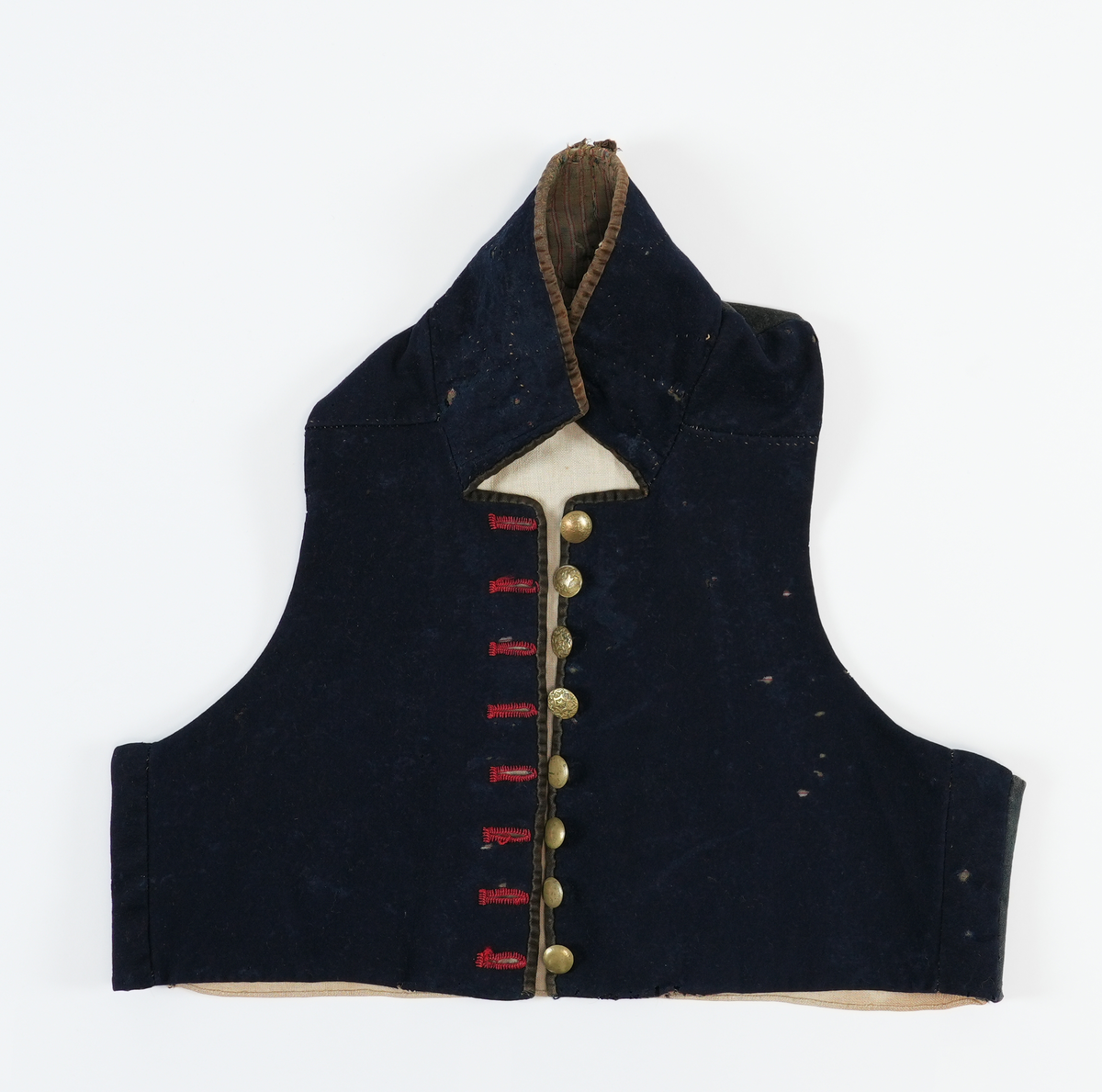 Vest sydd av mørkeblått klede med rygg av linlerret. Framstykka er fôra med stripet toskaft bomulssstoff i brunt, raudt og grått. På høgre framstykke åtte knapphol sydde med raud kameltråd, på venstre åtte runde metallknappar med pålodda hemper. Knappane er støypte skillingsknappar, nokre graverte. Slitne. Høg ståkrage stukken med prikkesting av ubleika lingarn. Krage og opning kanta med brune (svarte?) flyelsband. Fôr bak knapper av blått ribbet ullstoff.
