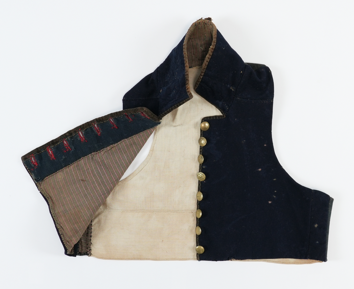 Vest sydd av mørkeblått klede med rygg av linlerret. Framstykka er fôra med stripet toskaft bomulssstoff i brunt, raudt og grått. På høgre framstykke åtte knapphol sydde med raud kameltråd, på venstre åtte runde metallknappar med pålodda hemper. Knappane er støypte skillingsknappar, nokre graverte. Slitne. Høg ståkrage stukken med prikkesting av ubleika lingarn. Krage og opning kanta med brune (svarte?) flyelsband. Fôr bak knapper av blått ribbet ullstoff.
