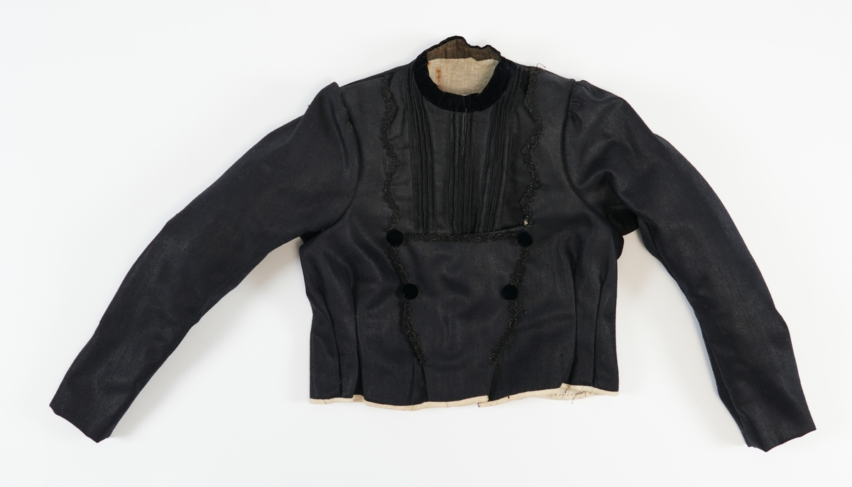 Svart jakke i ullstoff med kvitt linfôr. Dekorelement i form av biser (tre sett med fem bretter), fløyelstoff på kragen og fire falske knapper på framstykket. Svarte glassperlebånd er sydd som dekor rundt fronten på jakka og skjuler åpningen. To innsnitt på sidene av framstykket. Jakka festast saman av 17 hekter på høgre framstykket (påkledd) og fire på venstre skulder (påkledd). Opning midt framme som ein lukka med 12 metallhekter. For å skjule denne lukkinga bretta ein over dekorstykket. Nedre del av dekorstykket vert festa med fire metallhekter i venstre framstykket. Øvre del av dekorstykket vert festa med ei metallhekte, frå venstre til høgre. Nederste del av jakka har kvitt kanteband av lin. Jakka har puffermer. Bakstykket har fire biser midt på ryggen. Ryggen er satt saman av tre stoffsykker som forma jakka etter kroppsfasong. 

Innsida av halsopningen er kanta med eit grønnbrunt band av bomull (?). Heile jakka er sydd med maskin, men med noko hansaum på detaljane. Saumane på innsida ber preg av å ha blitt sydd ut slik at jakka vert større. 