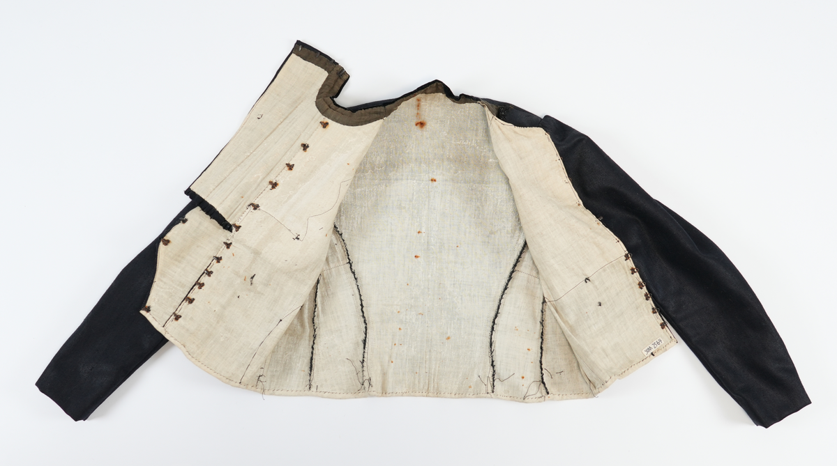 Svart jakke i ullstoff med kvitt linfôr. Dekorelement i form av biser (tre sett med fem bretter), fløyelstoff på kragen og fire falske knapper på framstykket. Svarte glassperlebånd er sydd som dekor rundt fronten på jakka og skjuler åpningen. To innsnitt på sidene av framstykket. Jakka festast saman av 17 hekter på høgre framstykket (påkledd) og fire på venstre skulder (påkledd). Opning midt framme som ein lukka med 12 metallhekter. For å skjule denne lukkinga bretta ein over dekorstykket. Nedre del av dekorstykket vert festa med fire metallhekter i venstre framstykket. Øvre del av dekorstykket vert festa med ei metallhekte, frå venstre til høgre. Nederste del av jakka har kvitt kanteband av lin. Jakka har puffermer. Bakstykket har fire biser midt på ryggen. Ryggen er satt saman av tre stoffsykker som forma jakka etter kroppsfasong. 

Innsida av halsopningen er kanta med eit grønnbrunt band av bomull (?). Heile jakka er sydd med maskin, men med noko hansaum på detaljane. Saumane på innsida ber preg av å ha blitt sydd ut slik at jakka vert større. 