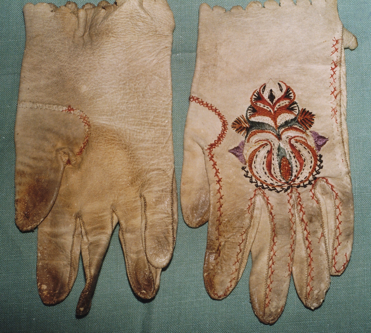 Ett par handskar av vitt skinn med uddstansad krage, broderi på handens ovansida i rött, grönt, svart, vitt och lila silke i plattsöm, stjälksöm och flätsöm, motiv av lökform med blad, flätsöm kring tumme och fingrar i rött. 

Birgitta Blixt 2022