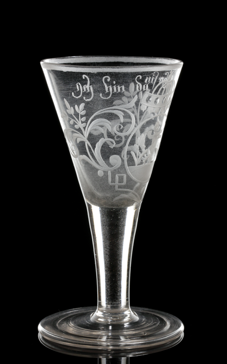 Vinglas av spetsglasmodell med konisk kupa som övergår i ett draget ben, Graverat motiv i form av en medaljon gm ed en amorin som sträcker sig efter ett bevingat hjärta, och över detta något som troligen är en krona. Motivet flankeras av växtmotiv, samt texten "Geh hin.... du falseher sin" som är placerad under mynningen.
Inskrivet i huvudkatalogen 1964.