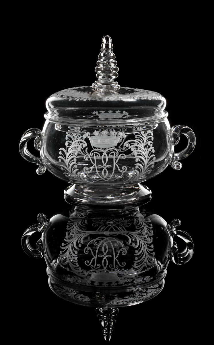 Smörask bestående av skål med två hänklar och lock med profilerad knopp.
På vardera motsatt sida graverade monogram med initialerna "M.P" och "E.K" under grevlig krona, flankerade av palm- och bärkvistar.
Bladranka på locket.
Inskriven i huvudkatalogen efter mars 1997.

Litteratur:
Se Seitz "Äldre Svenska glas", bild 51, Skånska glasbruket