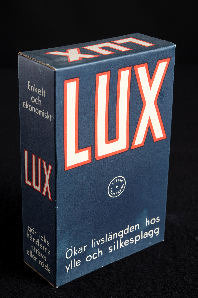 Förpackning av papp i mörkblå färg med text i vitt och rött. Formen är rektangulär, stående på höjden. Har innehållit tvålflingor, LUX Tvålflingor.