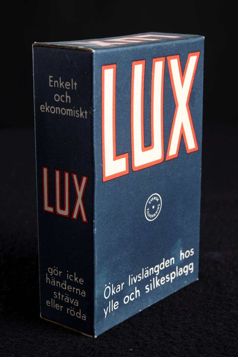 Förpackning av papp i mörkblå färg med text i vitt och rött. Formen är rektangulär, stående på höjden. Har innehållit tvålflingor, LUX Tvålflingor.