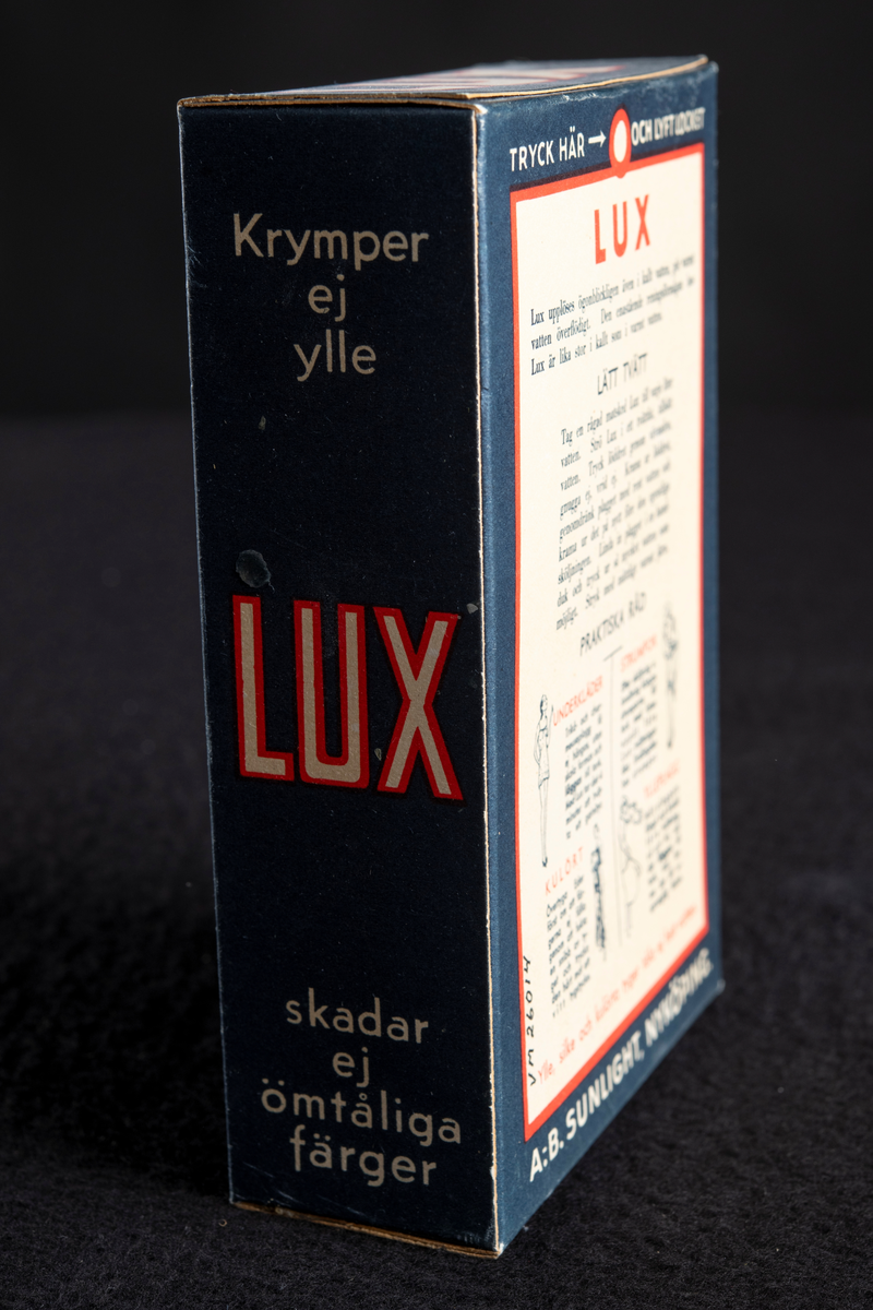 Förpackning av papp i mörkblå färg med text i vitt och rött. Formen är rektangulär, stående på höjden. Har innehållit tvålflingor, LUX Tvålflingor.
