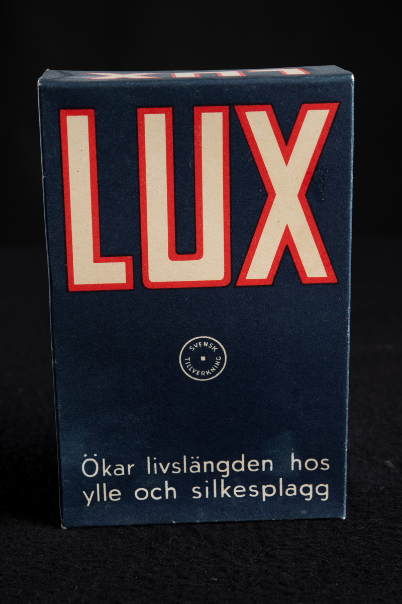 Förpackning av papp i mörkblå färg med text i vitt och rött. Formen är rektangulär, stående på höjden. Har innehållit tvålflingor, LUX Tvålflingor.