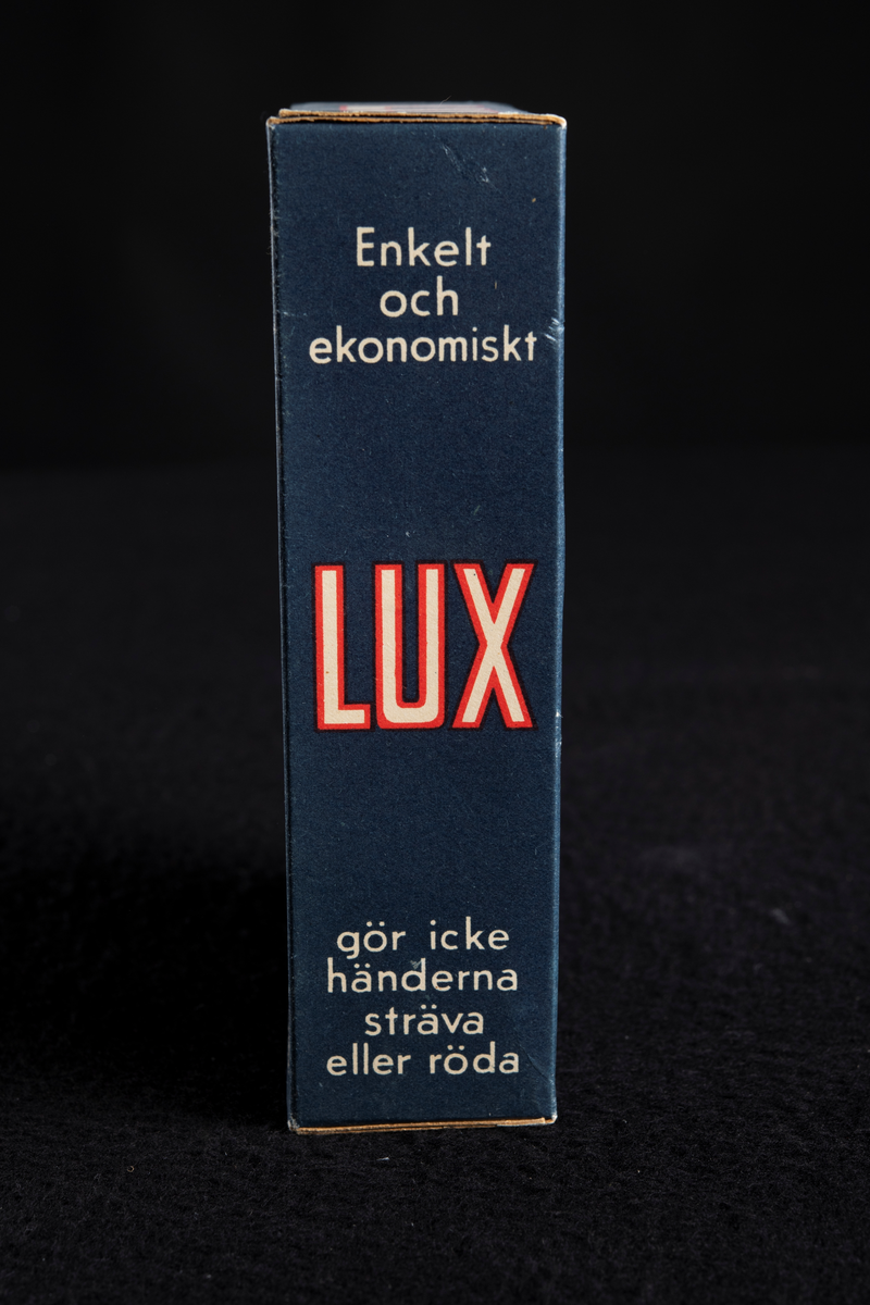 Förpackning av papp i mörkblå färg med text i vitt och rött. Formen är rektangulär, stående på höjden. Har innehållit tvålflingor, LUX Tvålflingor.