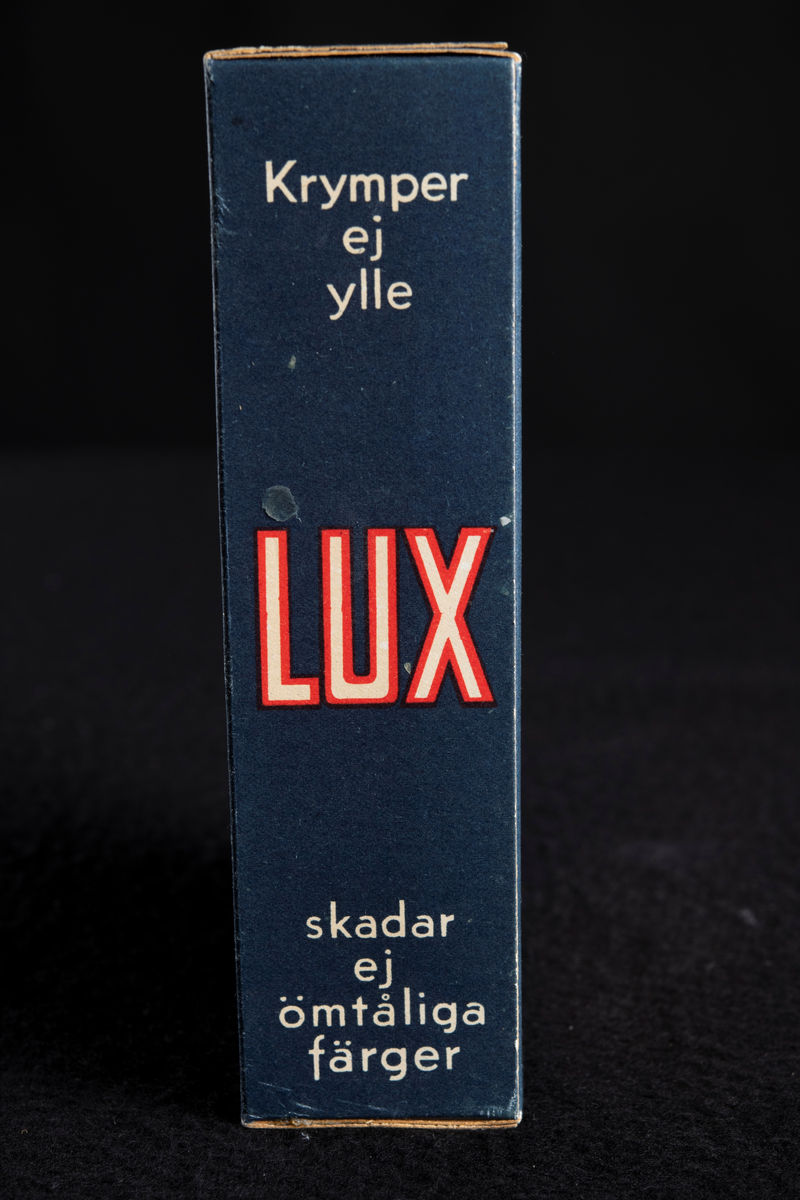 Förpackning av papp i mörkblå färg med text i vitt och rött. Formen är rektangulär, stående på höjden. Har innehållit tvålflingor, LUX Tvålflingor.