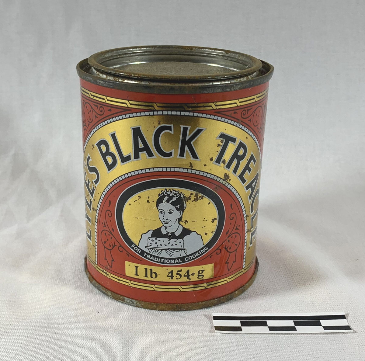 Metallboks med Lyle's Black Treacle, et britisk produkt brukt i tradisjonell matlaging. Boksen har en kapasitet på 1 lb (454 g) og er dekorert med røde, gule og svarte farger, med et ikonisk bilde av en kvinne som holder en kake, under slagordet "For Traditional Cooking." Boksen er sylinderformet og har et lokk som sannsynligvis har vært brukt til å forsegle det søte og mørke sirupsproduktet, kjent som treacle, som ofte brukes i baking.
