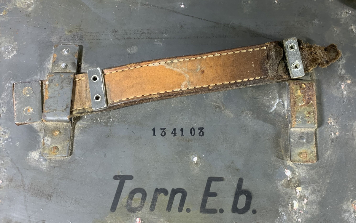 Boks til oppbevaring av tilbehør til en tysk radiomottaker, merket "Torn. E. b" (Tornister Empfänger b), en bærbar radio brukt av det tyske militæret under andre verdenskrig. Esken er merket med serienummeret "134103" og har rester av originalmaling, som viser slitasje og rust på flere områder. Det er et bærehåndtak av lær på siden, samt en låsemekanisme for å sikre innholdet. Lokket har en liten ventil, merket med "Auf" og "Zu" (åpen/lukket).