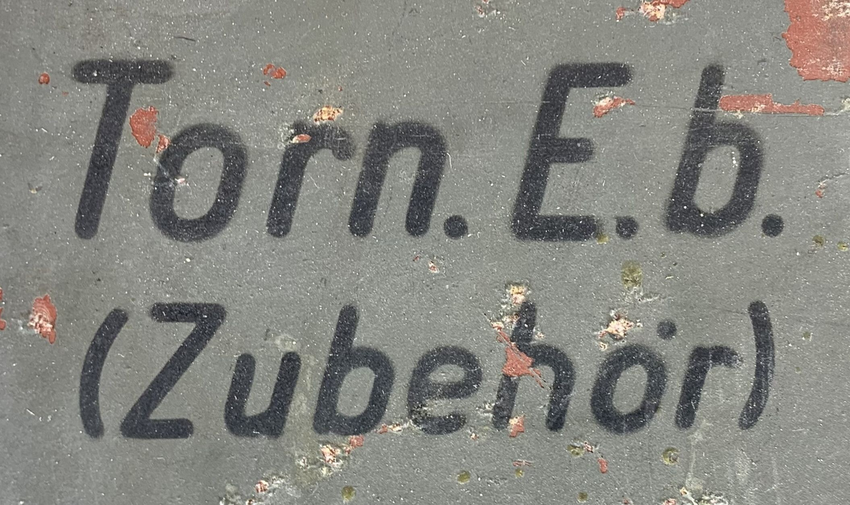 Boks til oppbevaring av tilbehør til en tysk radiomottaker, merket "Torn. E. b" (Tornister Empfänger b), en bærbar radio brukt av det tyske militæret under andre verdenskrig. Esken er merket med serienummeret "134103" og har rester av originalmaling, som viser slitasje og rust på flere områder. Det er et bærehåndtak av lær på siden, samt en låsemekanisme for å sikre innholdet. Lokket har en liten ventil, merket med "Auf" og "Zu" (åpen/lukket).