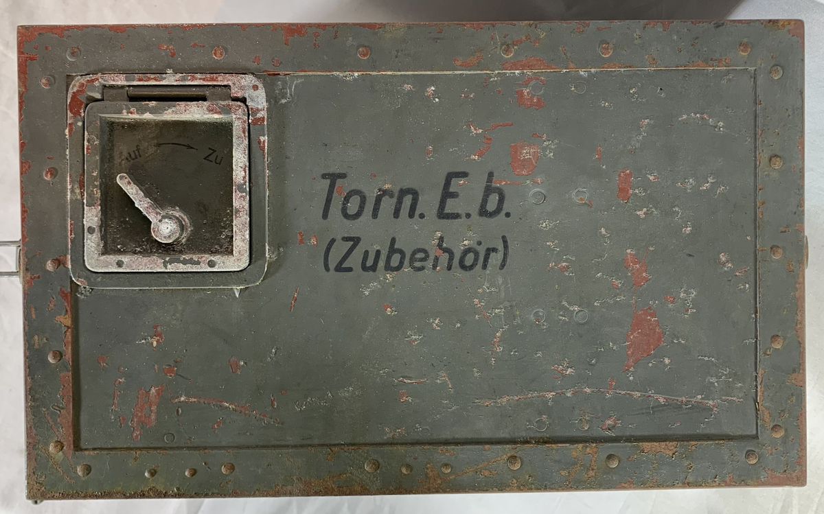 Boks til oppbevaring av tilbehør til en tysk radiomottaker, merket "Torn. E. b" (Tornister Empfänger b), en bærbar radio brukt av det tyske militæret under andre verdenskrig. Esken er merket med serienummeret "134103" og har rester av originalmaling, som viser slitasje og rust på flere områder. Det er et bærehåndtak av lær på siden, samt en låsemekanisme for å sikre innholdet. Lokket har en liten ventil, merket med "Auf" og "Zu" (åpen/lukket).