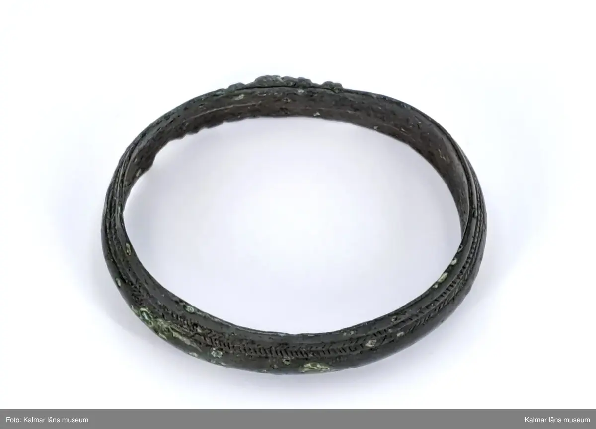 KLM 6059. Armring av brons. Nästan jämnbred och sluten. Smala åsar längs med ringen, två på var sida, är snett streckade tätt intill varandra med 12-13 tvärsträck växelvis, imiterande en omvirning.