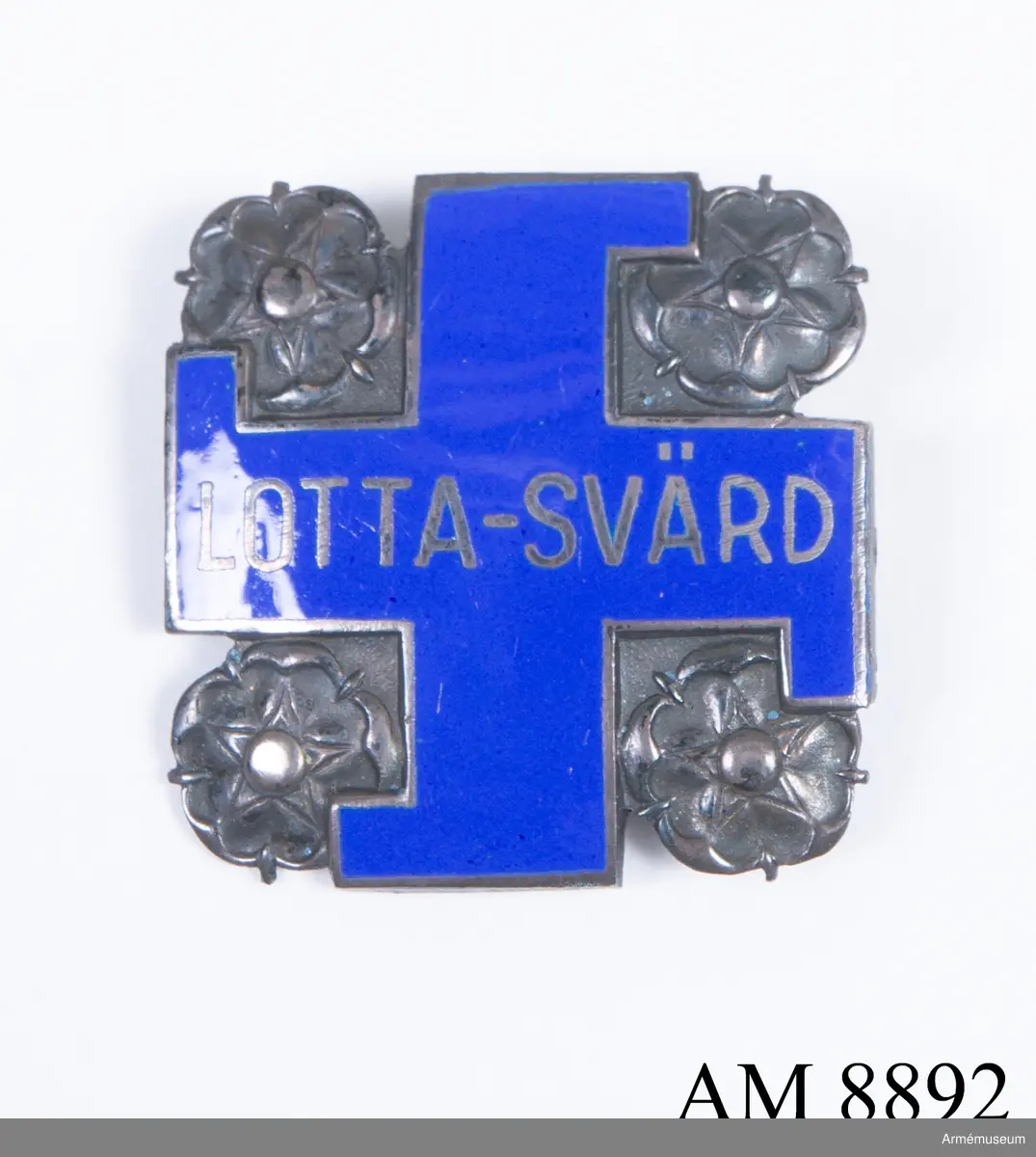 Av silver. Visar ett svastikakors i blå emalj med silverkant och namnet LOTTA-SVÄRD. Korset är omgivet av rosor i silver. Frånsidan bär silverstämplar och präglat nummer 63404.