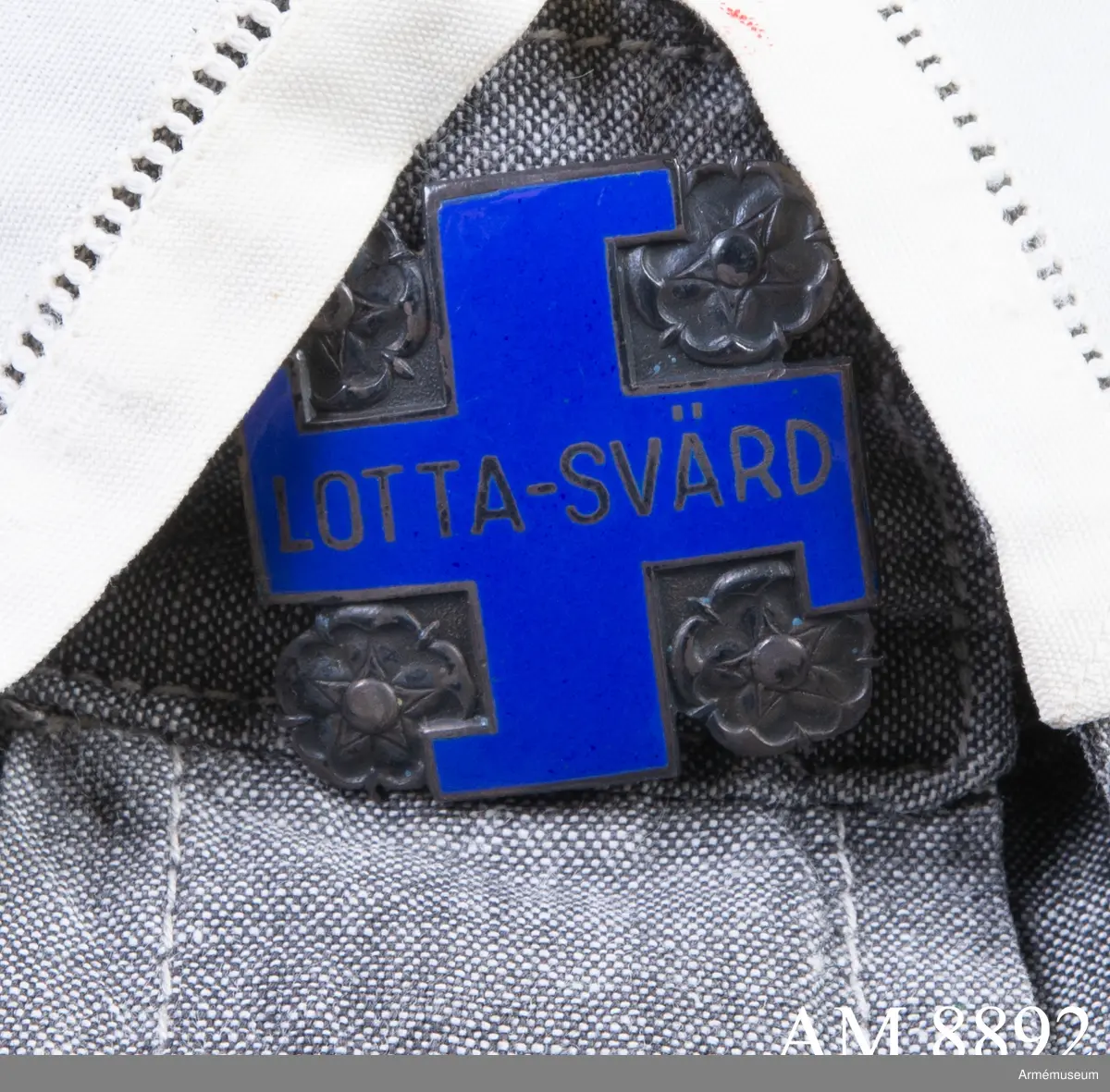 Av silver. Visar ett svastikakors i blå emalj med silverkant och namnet LOTTA-SVÄRD. Korset är omgivet av rosor i silver. Frånsidan bär silverstämplar och präglat nummer 63404.