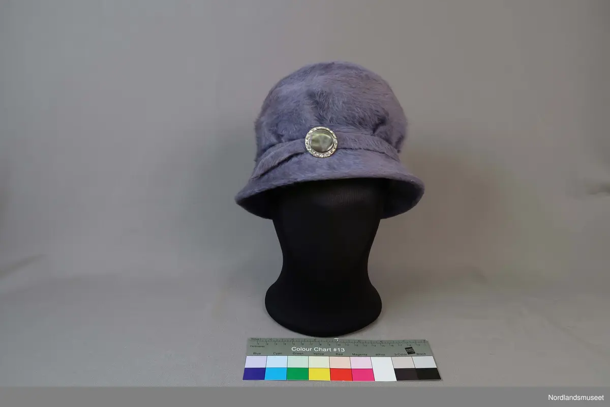 Lys lilla bøttehatt i børstet filt til dame. Bånd i samme materiale går rundt pullen og er festet med en pynteknapp. Hattebånd på innside i bomullslerret. Strikk er festet inni for å holde hatten på plass rundt frisyre.