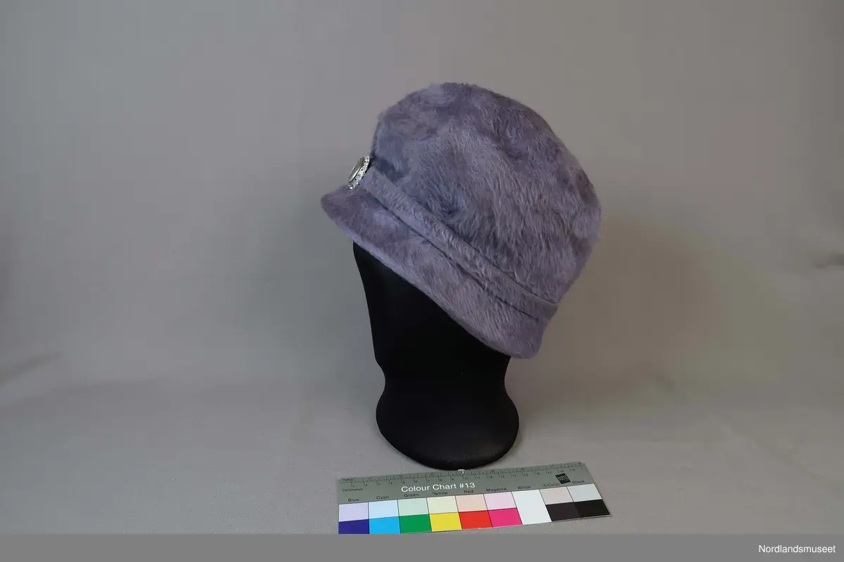 Lys lilla bøttehatt i børstet filt til dame. Bånd i samme materiale går rundt pullen og er festet med en pynteknapp. Hattebånd på innside i bomullslerret. Strikk er festet inni for å holde hatten på plass rundt frisyre.