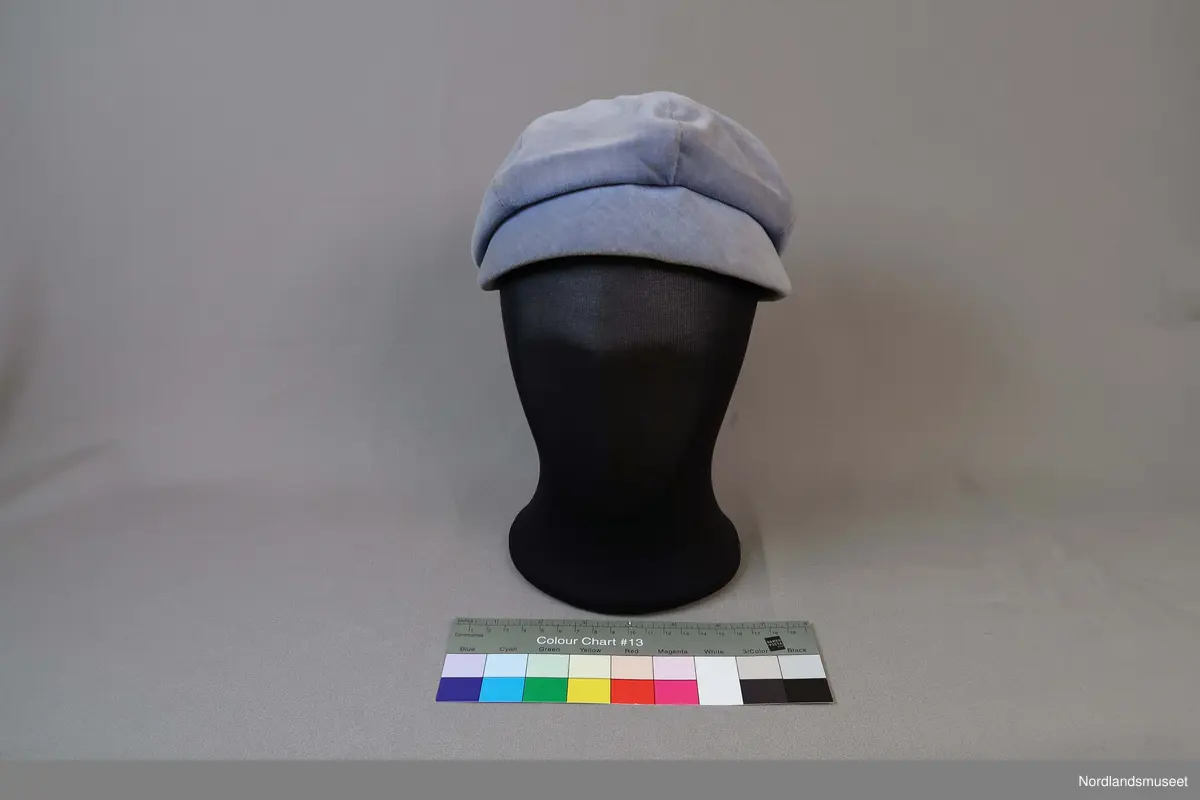 Liten hatt til dame i lys blå fløyel. Liten brem i forkant. Fôret med hvitt syntetisk stoff. Stoffkledd knapp øverst.