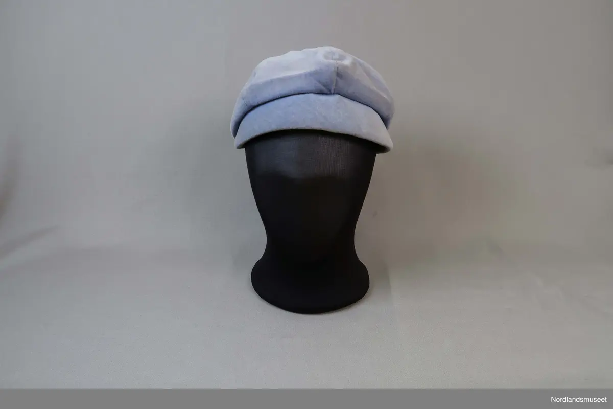 Liten hatt til dame i lys blå fløyel. Liten brem i forkant. Fôret med hvitt syntetisk stoff. Stoffkledd knapp øverst.