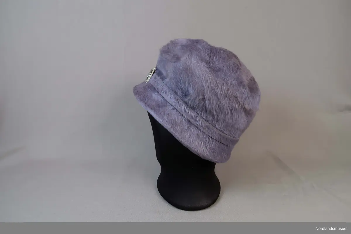 Lys lilla bøttehatt i børstet filt til dame. Bånd i samme materiale går rundt pullen og er festet med en pynteknapp. Hattebånd på innside i bomullslerret. Strikk er festet inni for å holde hatten på plass rundt frisyre.