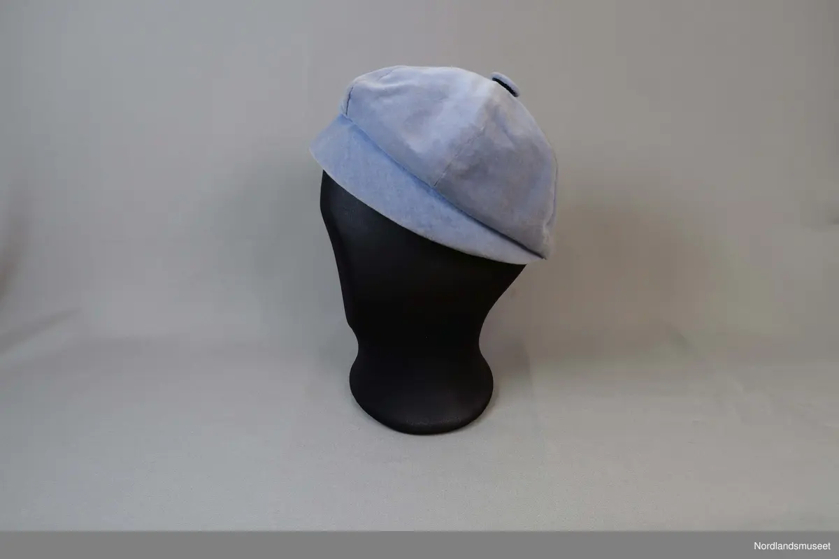 Liten hatt til dame i lys blå fløyel. Liten brem i forkant. Fôret med hvitt syntetisk stoff. Stoffkledd knapp øverst.