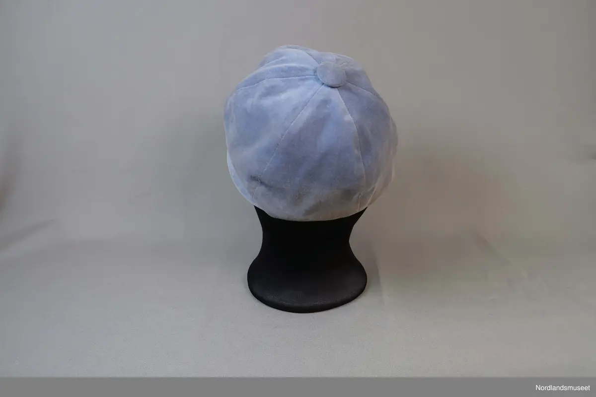 Liten hatt til dame i lys blå fløyel. Liten brem i forkant. Fôret med hvitt syntetisk stoff. Stoffkledd knapp øverst.